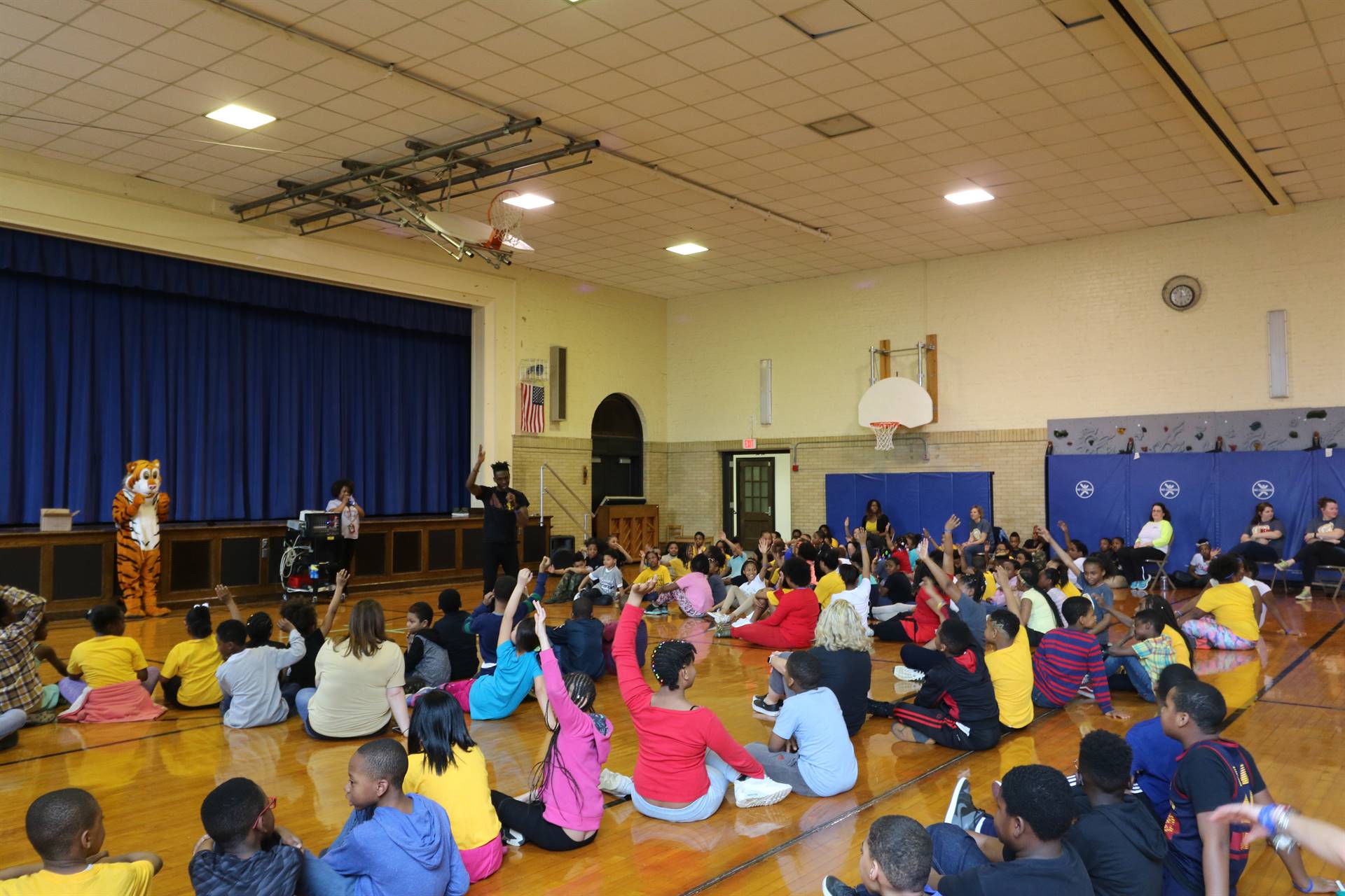 Students sit on gym floor, some with hands raised