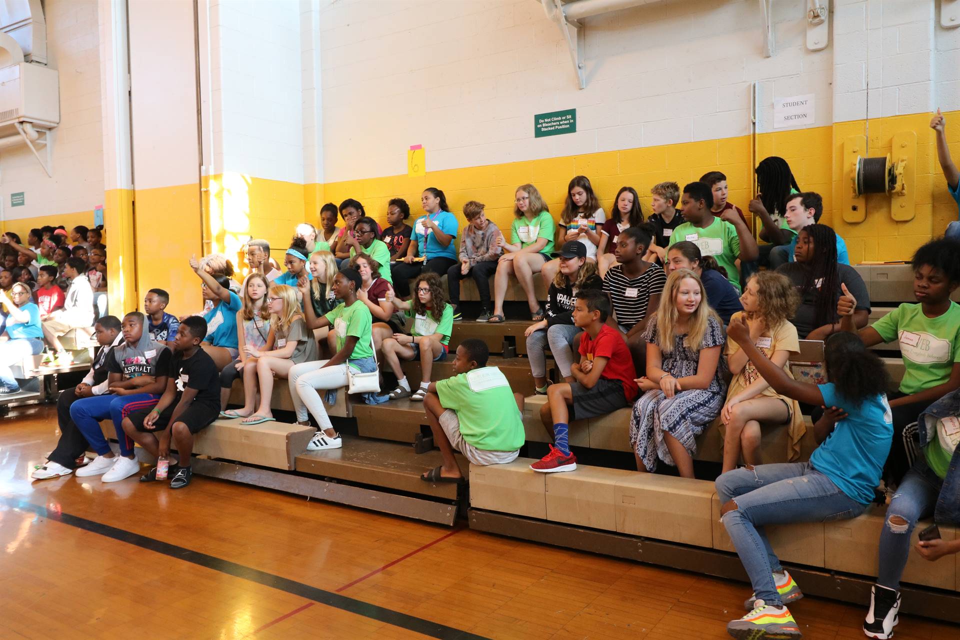 Students in gym bleachers with hands raised